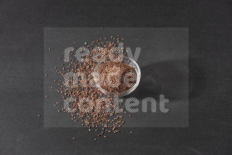 A glass bowl full of flaxseeds surrounded by the seeds on a black flooring