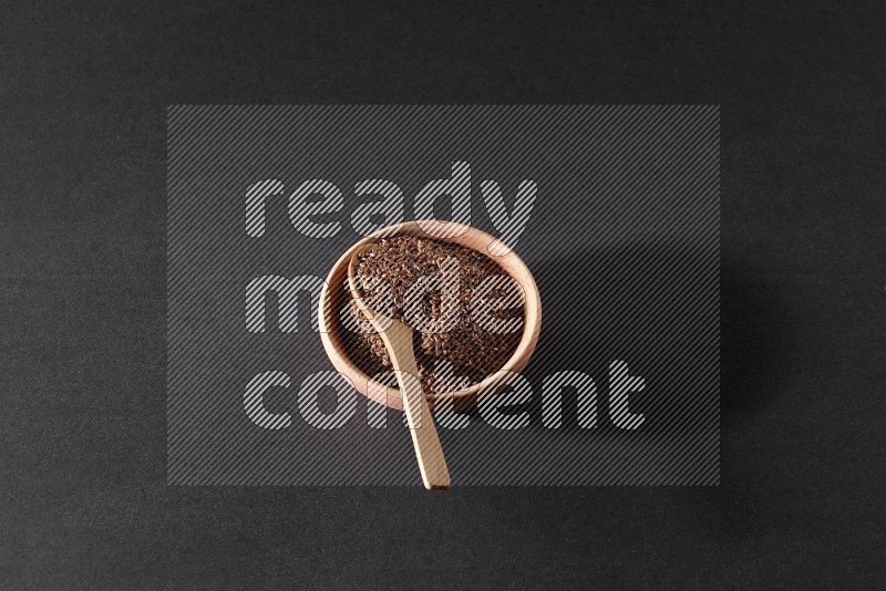A wooden bowl full of flaxseeds with wooden spoon full of the seeds on it on a black flooring