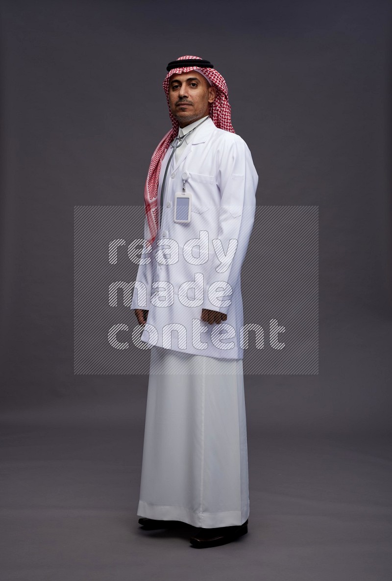 Saudi man wearing thob with lab coat and shomag with pocket employee badge with stethoscope standing interacting with the camera on gray background