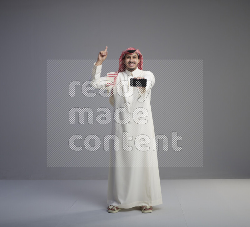 A Saudi man standing wearing thob and red shomag showing phone to camera on gray background