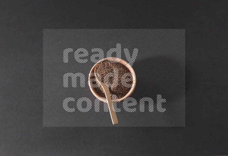 A wooden bowl full of flaxseeds with wooden spoon full of the seeds on it on a black flooring