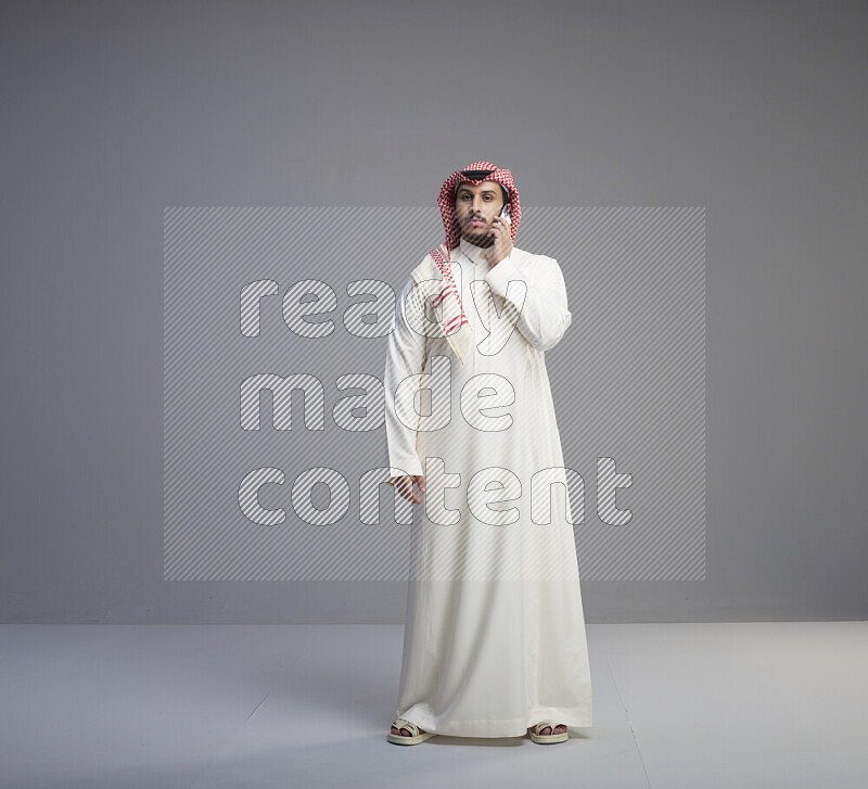 A Saudi man standing wearing thob and red shomag talking on phone on gray background