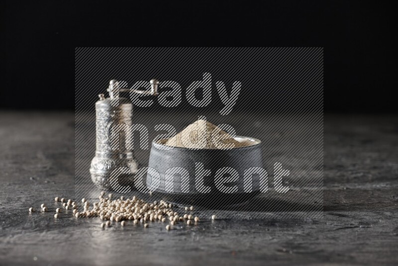 Black pottery bowl full of white pepper powder with pepper beads and metal pepper grinder on textured black flooring