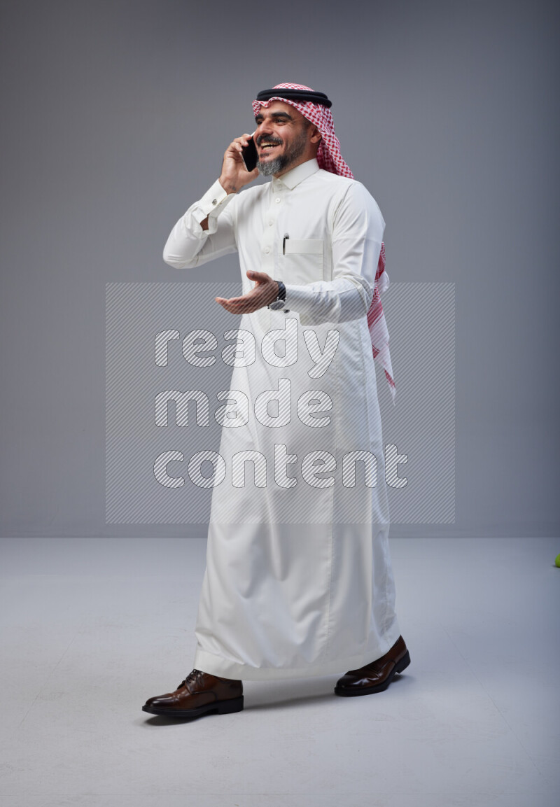 Saudi man Wearing Thob and red Shomag standing talking on phone on Gray background