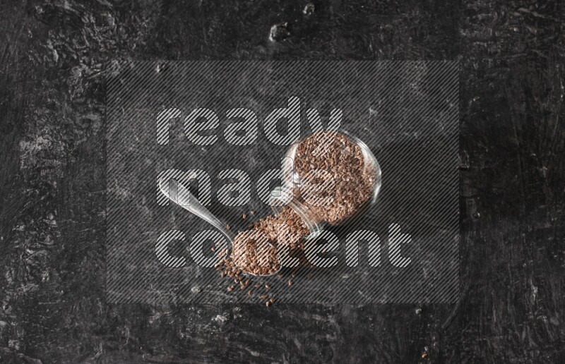 A glass spice jar full of flaxseeds flipped and seeds spread out with a metal spoon full of the seeds on a textured black flooring