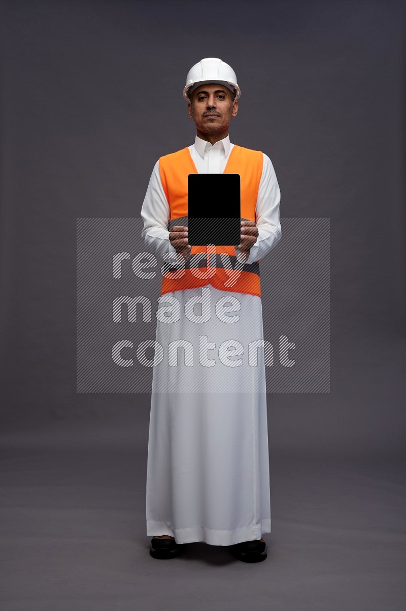 Saudi man wearing thob with engineer vest standing showing tablet to camera on gray background