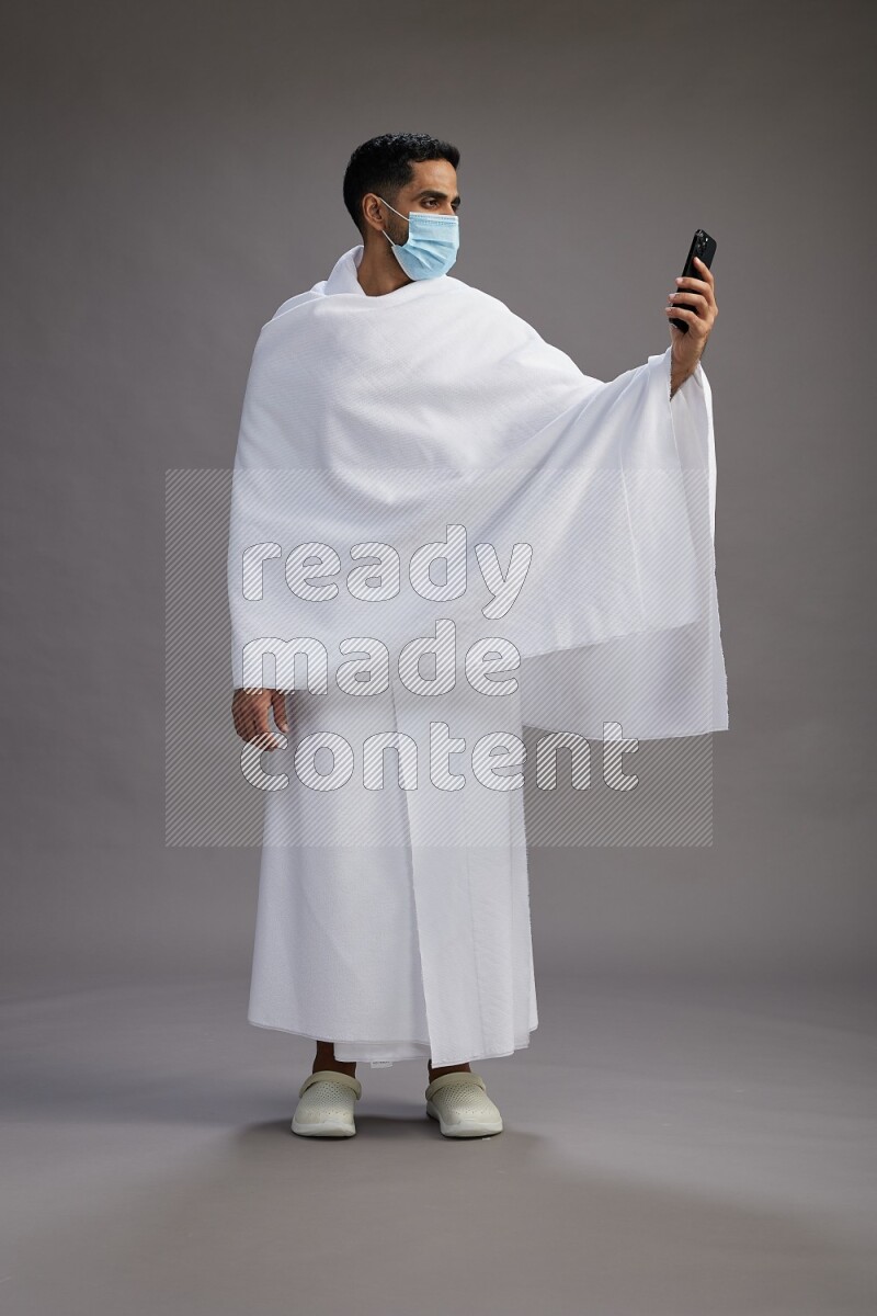 A man wearing Ehram with face mask Standing taking selfie on gray background