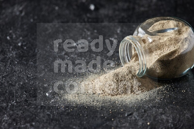 A flipped herbal glass jar full of white pepper powder with spilled powder on textured black flooring