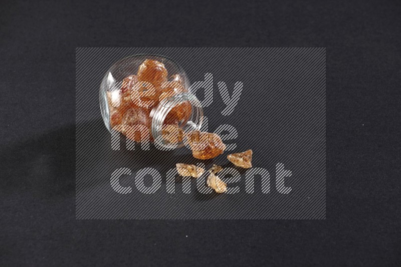 A glass spice jar full of gum arabic and jar is flipped with fallen pieces on black flooring