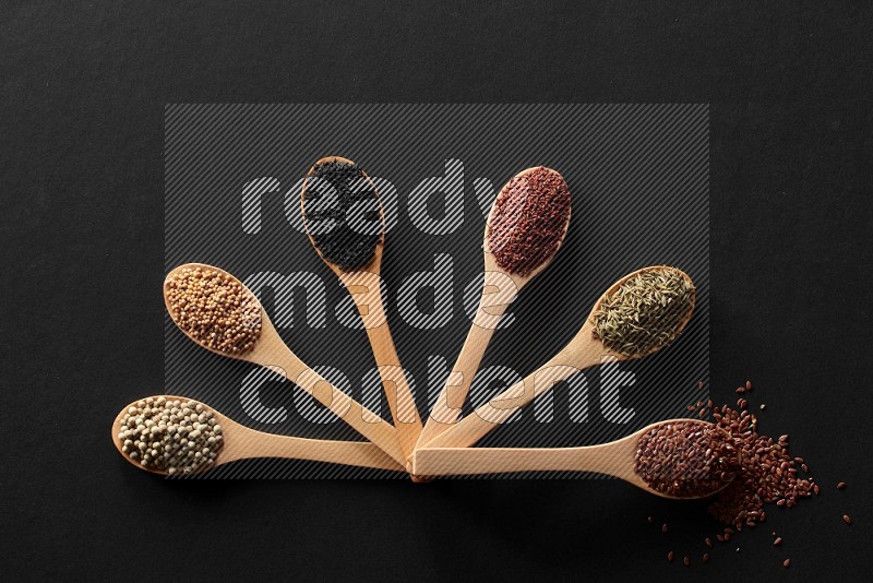 wooden spoons filled with white peppers, mustard seeds, black seeds, garden cress, cumin and flax on black flooring