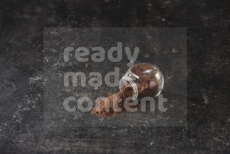 A flipped glass spice jar full of cloves powder and powder came out of it on textured black flooring