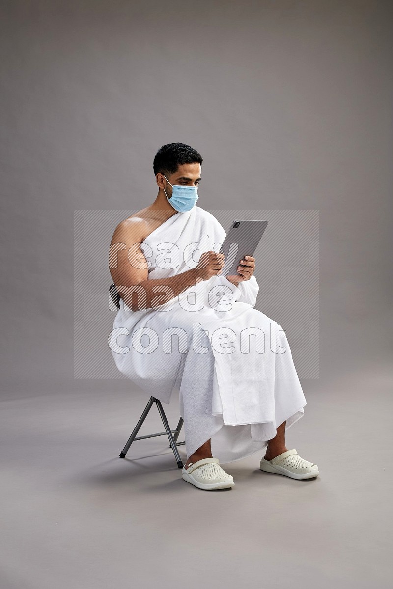 A man wearing Ehram with face mask sitting on chair holding a tablet on gray background