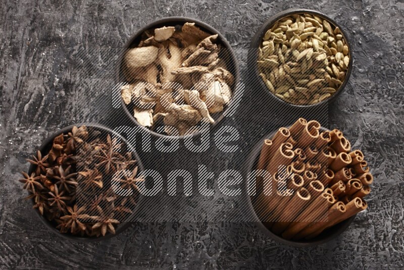 Ginger, Cardamom, Star anise and cinnamon sticks in 4 bowls on a textured black background
