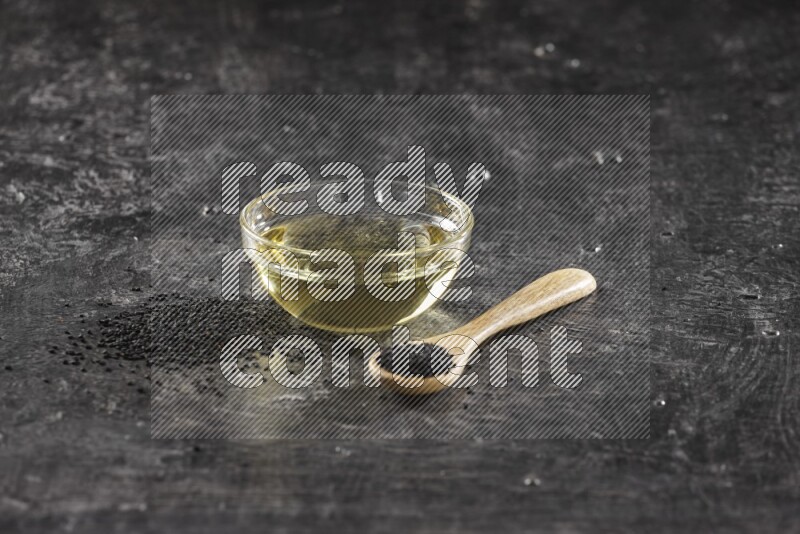 وعاء زجاجي ممتلئ بزيت حبة البركة وملعقة خشبية ممتلئة بالبذور مع بعض البذور متناثرة على أرضية سوداء