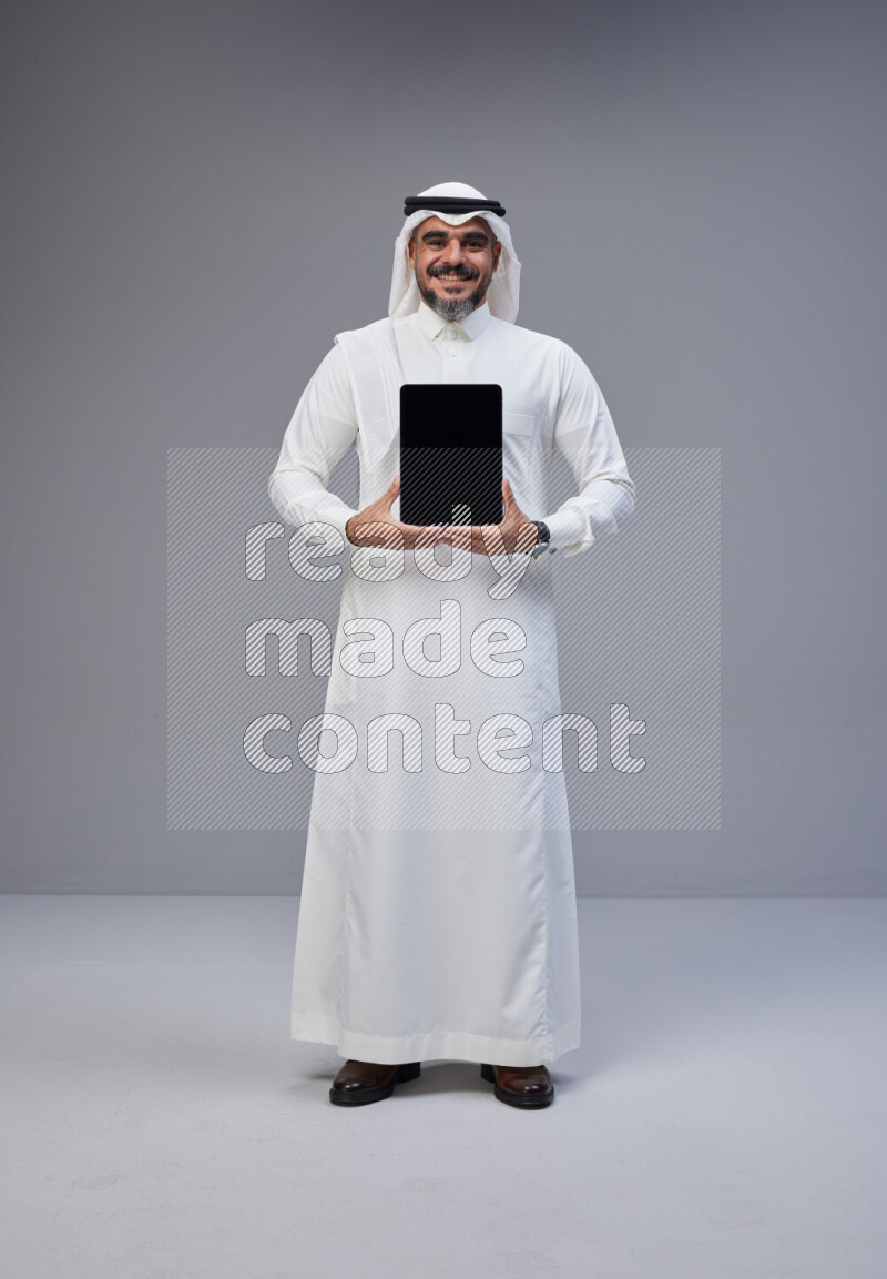 Saudi man Wearing Thob and white Shomag standing showing tablet to camera on Gray background