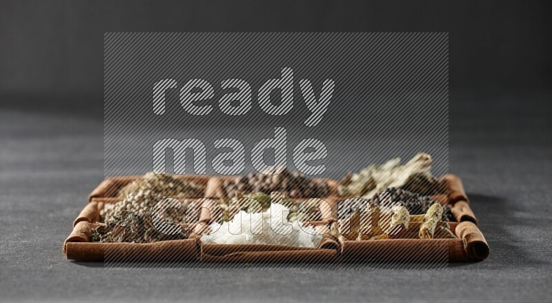 9 مربعات من أعواد القرفة ممتلئة بالهيل في المنتصف محاطة بأصابع الكركم والفلفل الأسود وورق الغار والبهار الحلو والكمون والفلفل الأبيض والريحان المجفف والملح على أرضية سوداء