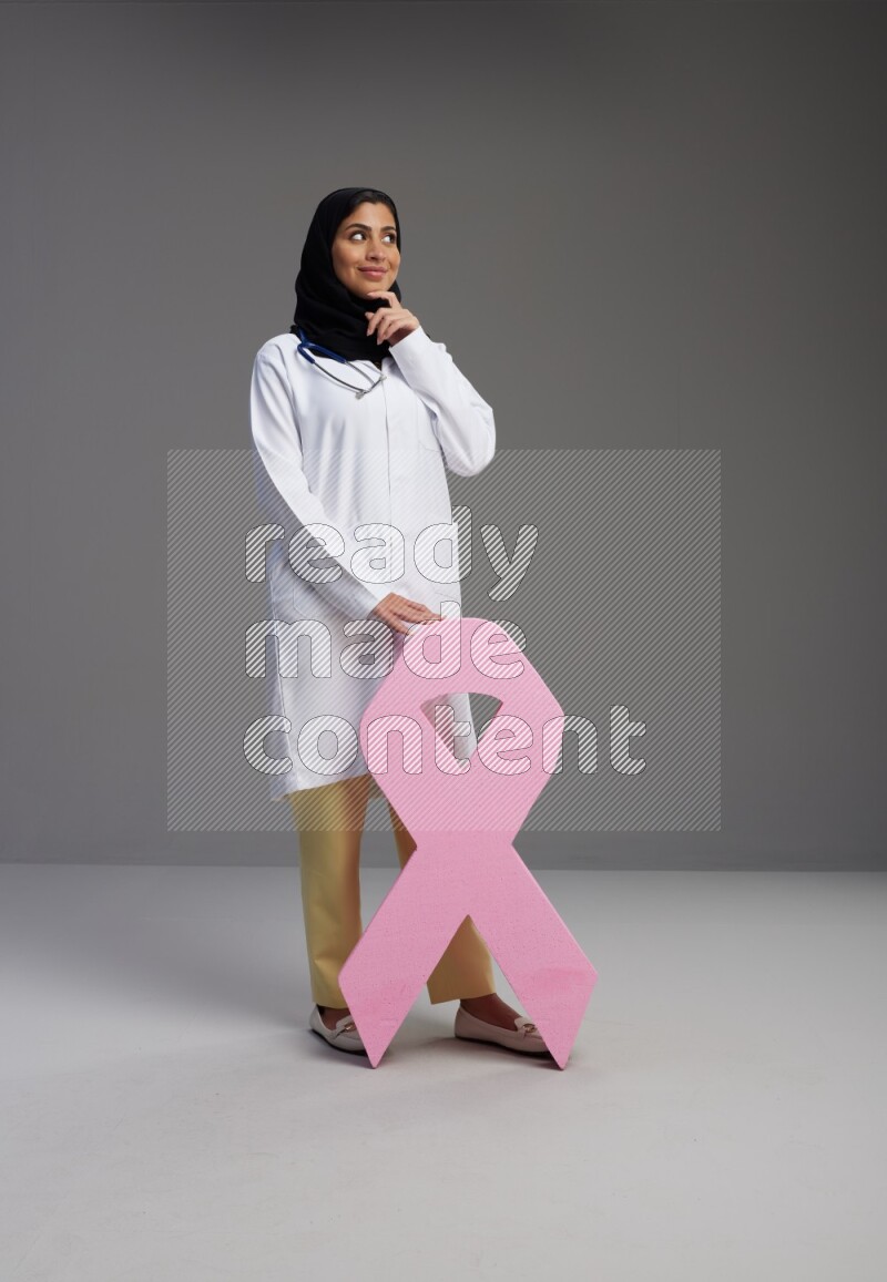 Saudi woman wearing lab coat with stethoscope standing holding awareness ribbon symbol standing on Gray background
