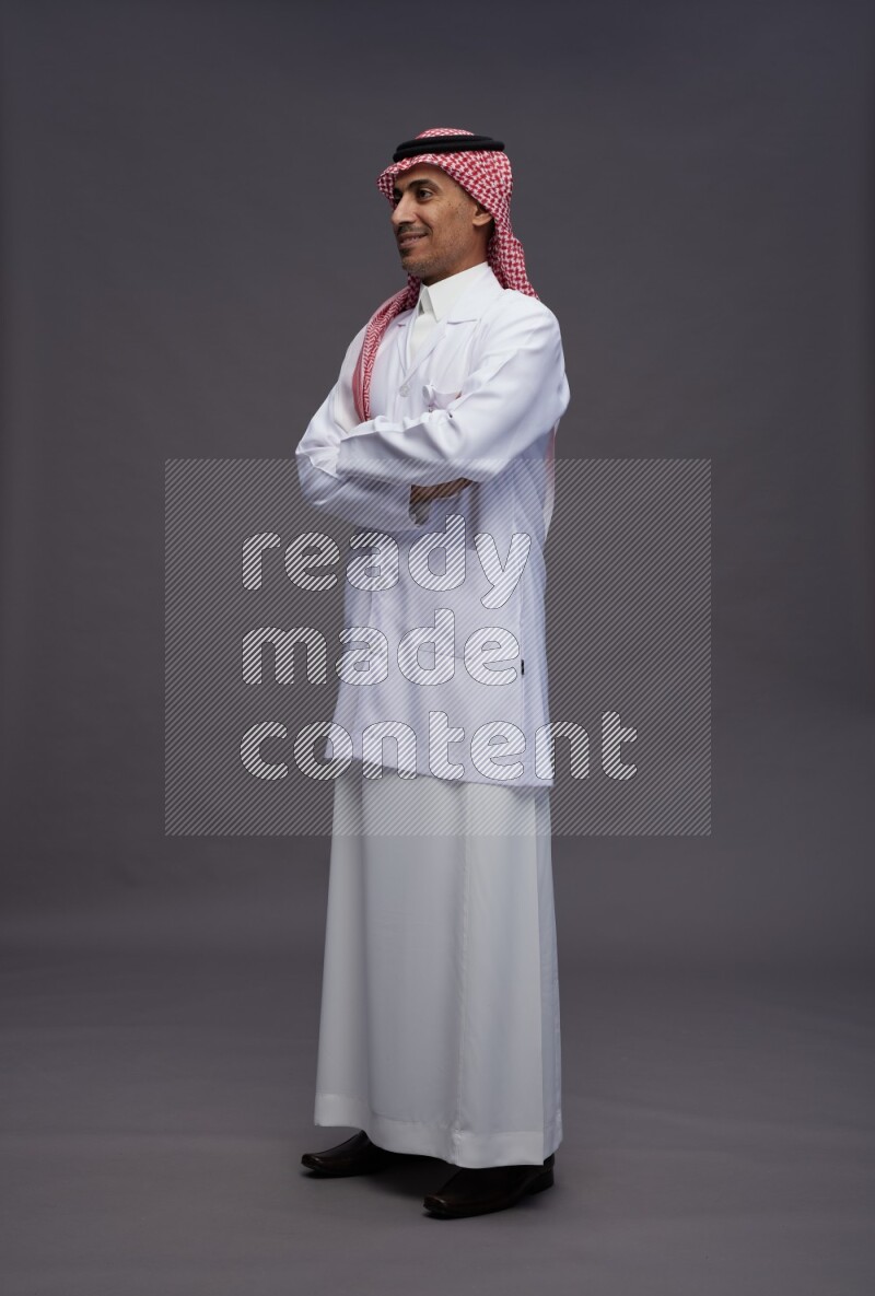 Saudi man wearing thob with lab coat and shomag with pocket employee badge standing with crossed arms on gray background