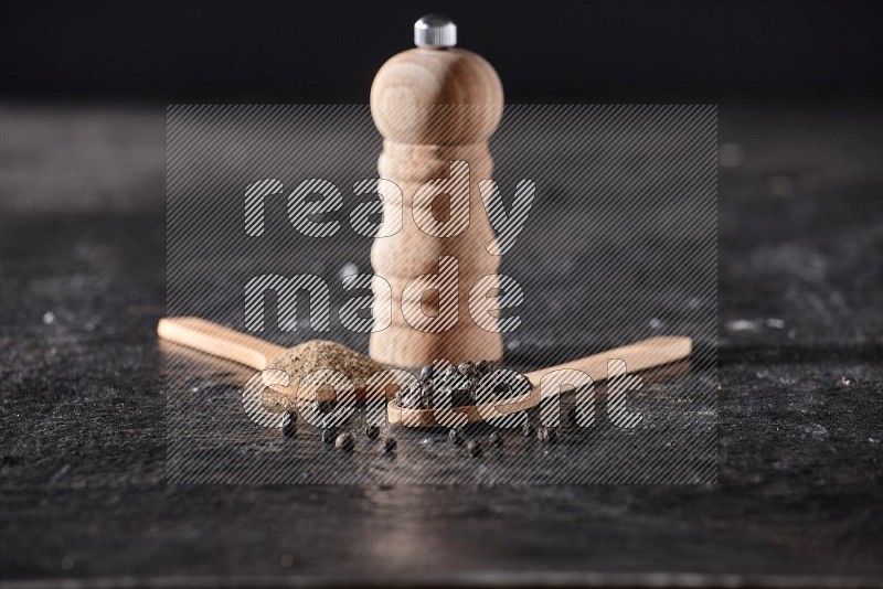 Black pepper powder and black pepper beads in wooden spoons and wooden grinder on a textured black flooring