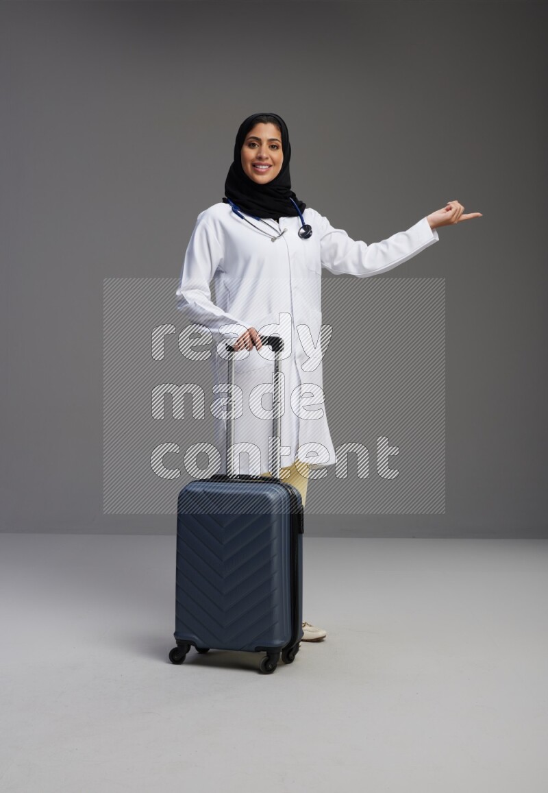 Saudi woman wearing lab coat with stethoscope standing holding Travel bag on Gray background