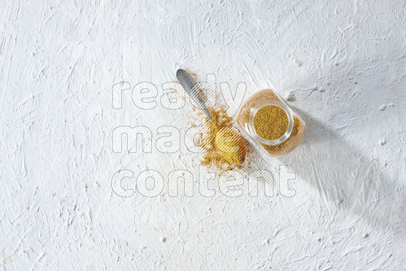 A glass spice jar and metal spoon full of turmeric powder on textured white flooring