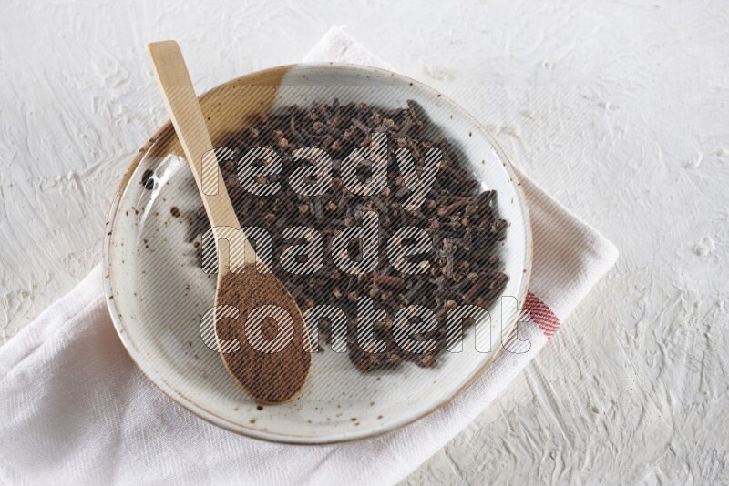 A Pottery plate full of whole cloves and a wooden spoon full of cloves powder in it on a textured white background