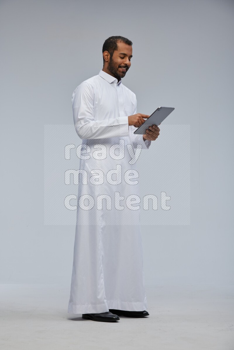 Saudi man wearing Thob standing working on tablet on Gray background