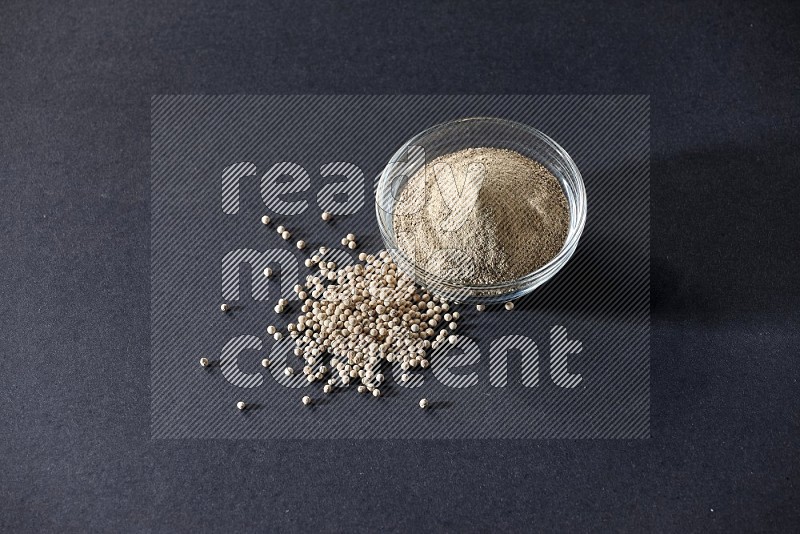 A glass bowl full of white pepper powder with white pepper beads on black flooring