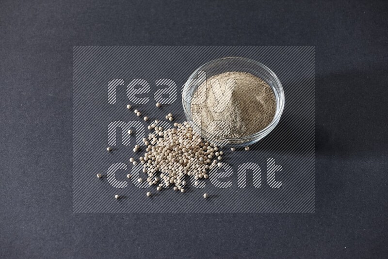 وعاء زجاجي مملوء بمسحوق الفلفل الأبيض مع حبات الفلفل الأبيض على أرضية سوداء