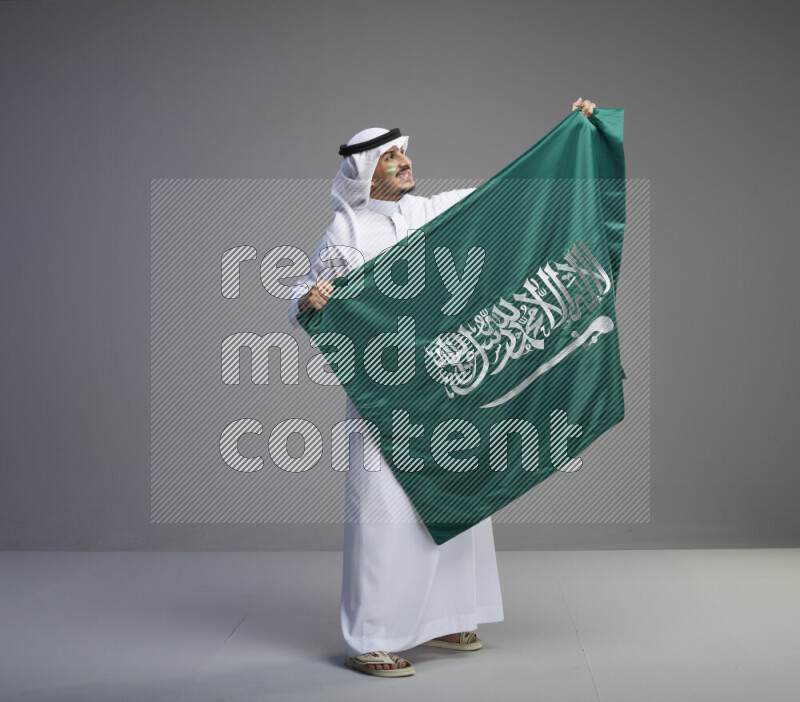 A Saudi man standing wearing thob and white shomag with face painting holding big Saudi flag on gray background