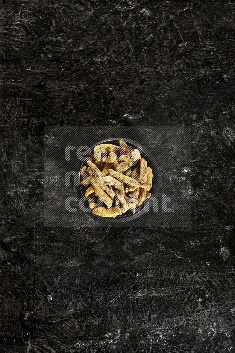 A black pottery bowl full of dried turmeric whole fingers on textured black flooring