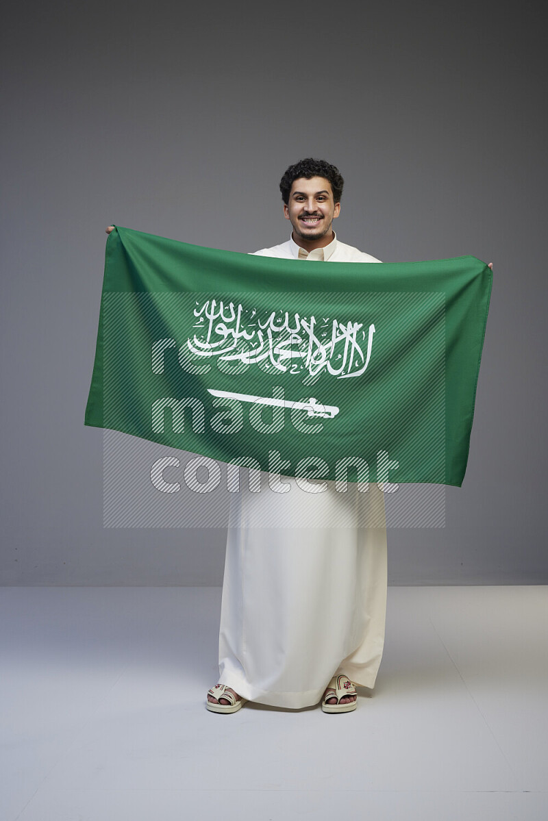 A Saudi man standing wearing thob holding big Saudi flag on gray background