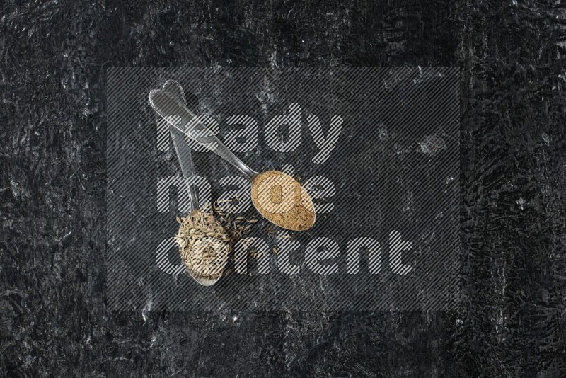 ملعقتان معدنيتان مملوءتان بحبوب الكمون وبودرة الكمون على أرضية سوداء