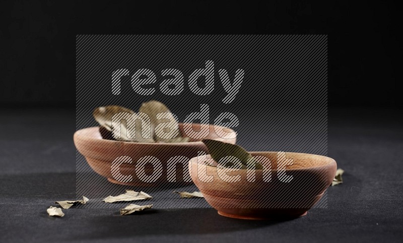 2 wooden bowls full of dried bay leaves with more leaves spread on black flooring