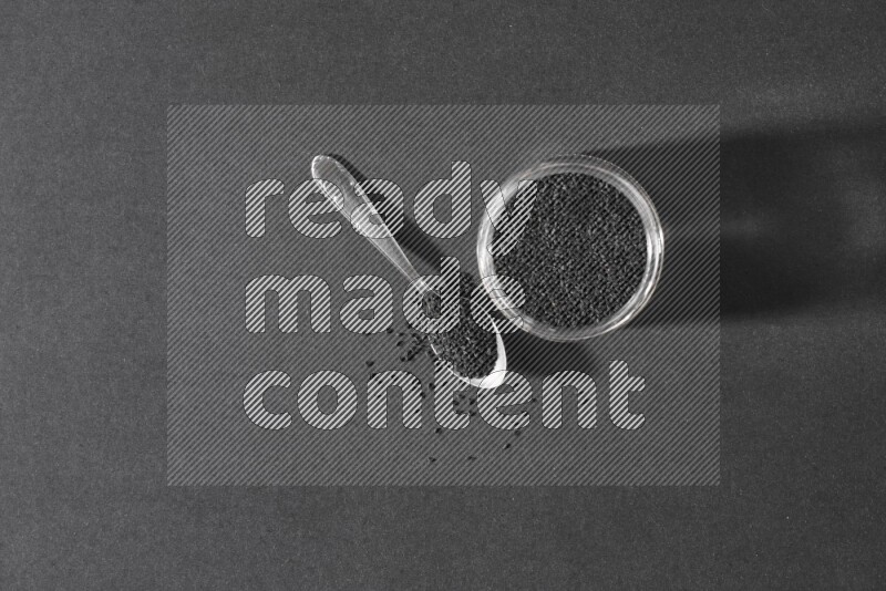 A glass jar full of black seeds with a metal spoon full of the seeds on a black flooring