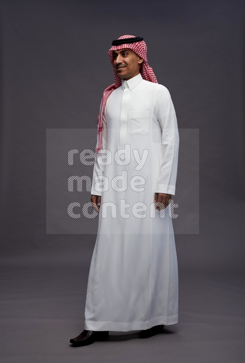 Saudi man wearing thob and shomag standing interacting with the camera on gray background