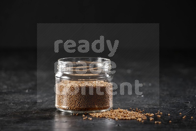 A glass jar full of mustard seeds and jar is flipped and seeds spread out on a textured black flooring