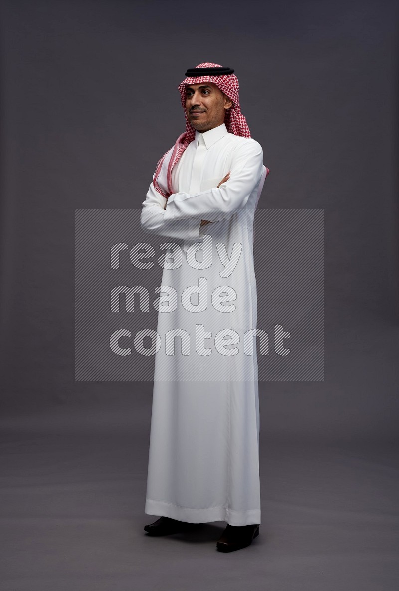 Saudi man wearing thob and shomag standing with crossed arms on gray background