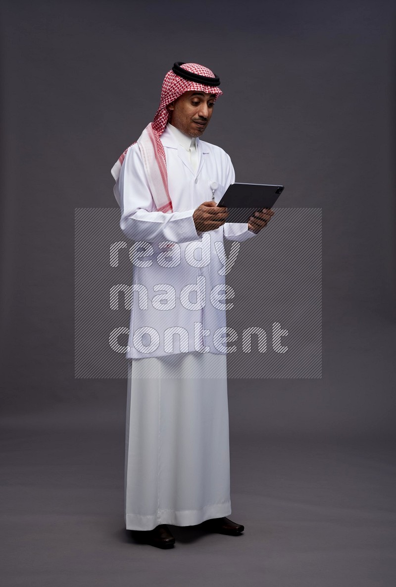 Saudi man wearing thob with lab coat and shomag with pocket employee badge standing working on tablet on gray background