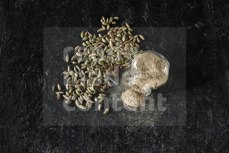 وعاء زجاجي للتوابل مقلوب وملعقة معدنية ممتلئة ببودرة الهيل وحبوب الهيل متناثرة على خلفية سوداء