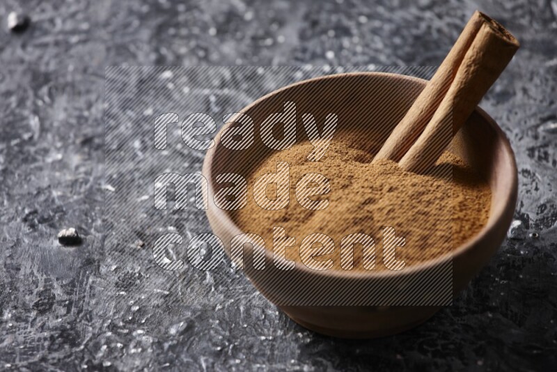 Wooden bowl full of cinnamon powder and a cinnamon stick on a textured black background