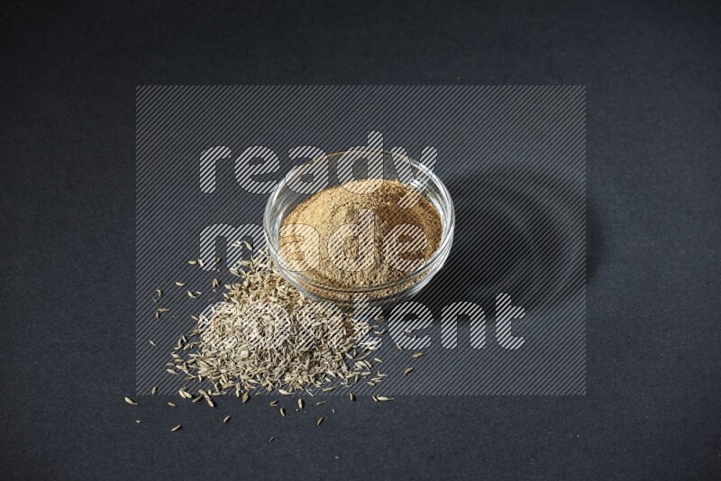 A glass bowl full of cumin powder with cumin seeds beside it on black flooring