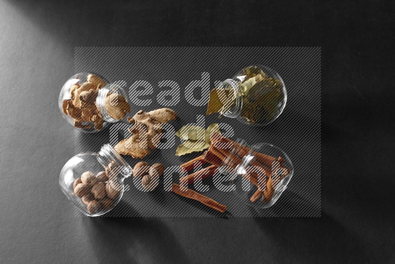4 glass spice jars full of laurel bay leaves, ginger, cinnamon and nutmeg on black flooring