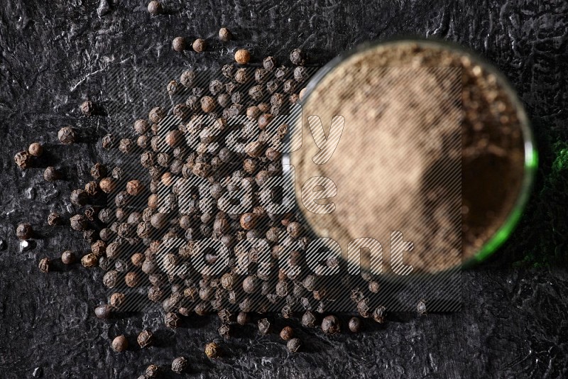 A glass cup full of black pepper powder with beads beside it on a textured black flooring