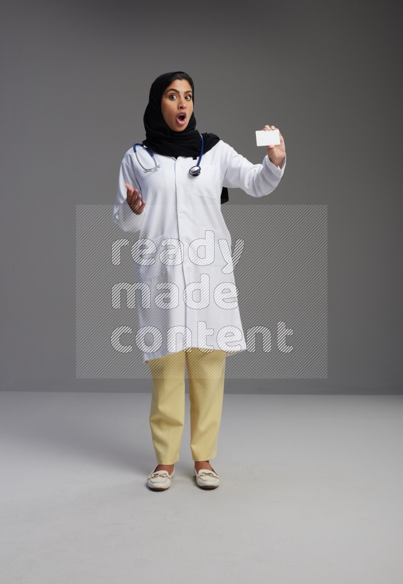 Saudi woman wearing lab coat with stethoscope standing holding ATM card on Gray background