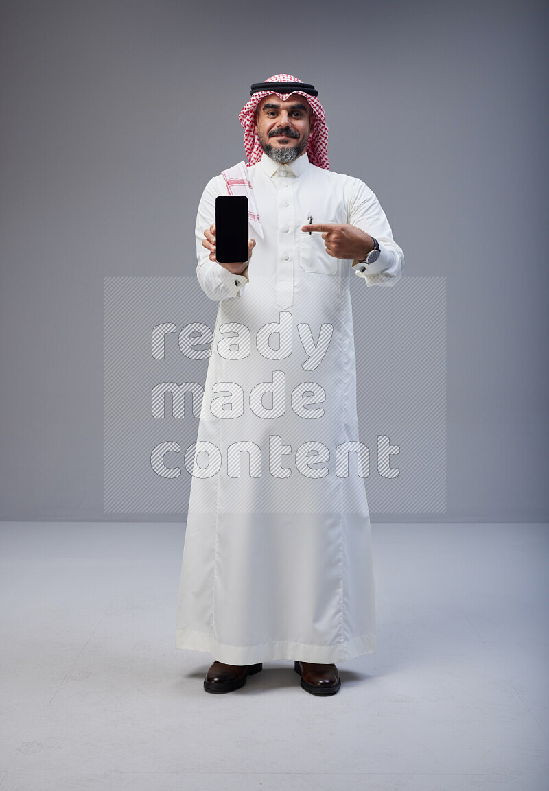 Saudi man Wearing Thob and red Shomag standing showing phone to camera on Gray background