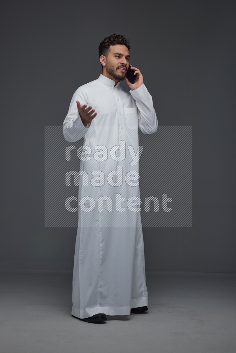 A Saudi man wearing Thobe and talking in the phone while standing and making different poses eye level on a gray background