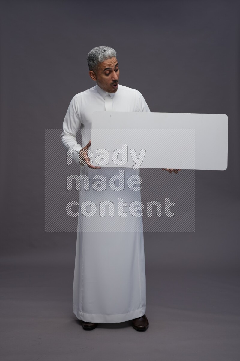 Saudi man wearing thob standing holding board on gray background