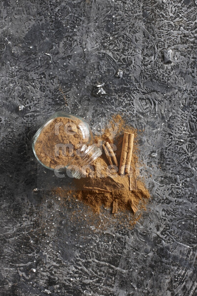 Flipped glass herbs jar full of cinnamon powder with cracked cinnamon sticks on a textured black background