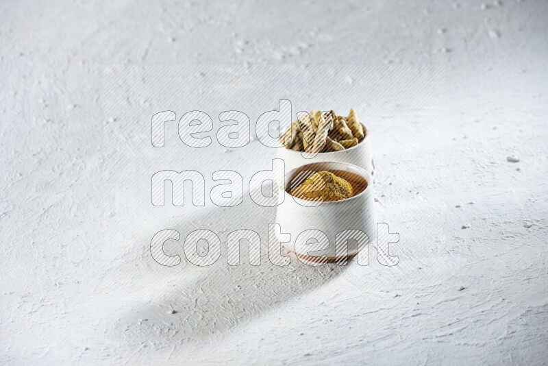 2 beige pottery bowl full of turmeric powder and dried turmeric whole fingers on textured white flooring